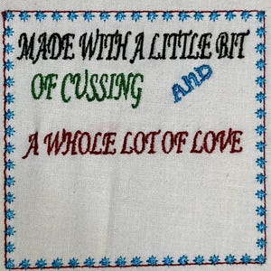 May include: White fabric with a blue, red, green, and black embroidered border. The text reads "Made with a little bit of cussing and a whole lot of love."