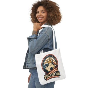 May include: White tote bag featuring a soccer ball with a crown, the American flag, and the text "World Cup 2026 Canada USA Mexico". The bag has white straps and is being held by a person wearing a denim jacket.
