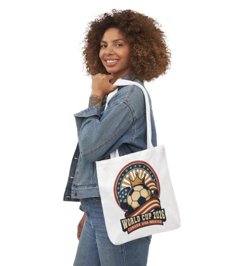 May include: White tote bag featuring a soccer ball with a crown, the American flag, and the text "World Cup 2026 Canada USA Mexico". The bag has white straps and is being held by a person wearing a denim jacket.
