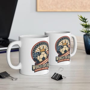 May include: Two white ceramic mugs with a soccer-themed design. The design features a soccer ball with a crown, surrounded by a red, white, and blue circular emblem with the text "WORLD CUP 2026 CANADA USA MEXICO".