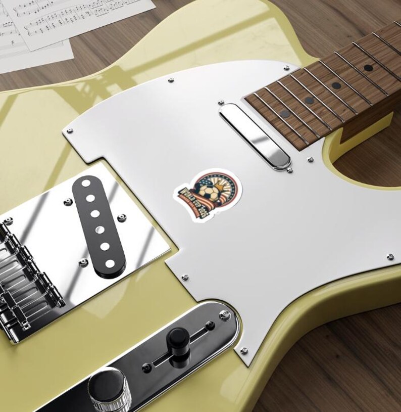 May include: A sticker with a soccer ball design, featuring the text "World Cup 2026". The sticker has a vintage aesthetic with red, white, and blue colors, and is placed on a light yellow electric guitar.
