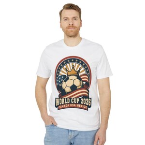 May include: White t-shirt featuring a soccer ball with a crown, set against a backdrop of stars and stripes. The design includes the text "WORLD CUP 2026 CANADA USA MEXICO". The shirt is worn by a person.