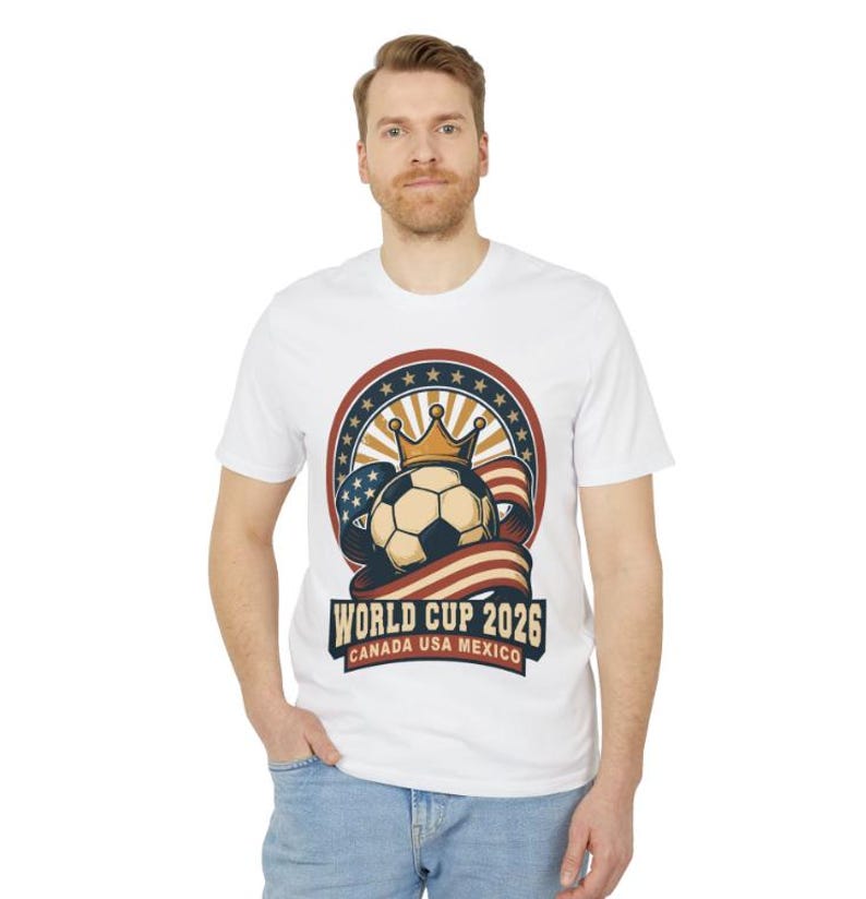 May include: White t-shirt featuring a soccer ball with a crown, set against a backdrop of stars and stripes. The design includes the text "WORLD CUP 2026 CANADA USA MEXICO". The shirt is worn by a person.