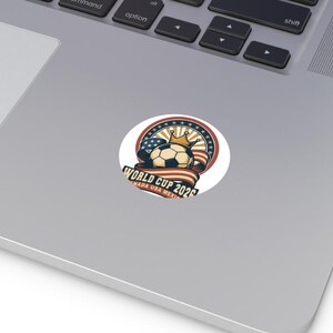 May include: A round sticker featuring a soccer ball wearing a gold crown, set against a backdrop of a red, white, and blue ribbon and stars. The text "WORLD CUP 2026 CANADA USA MEXICO" is displayed.