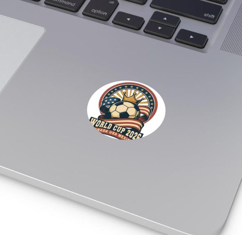 May include: A round sticker featuring a soccer ball wearing a gold crown, set against a backdrop of a red, white, and blue ribbon and stars. The text "WORLD CUP 2026 CANADA USA MEXICO" is displayed.