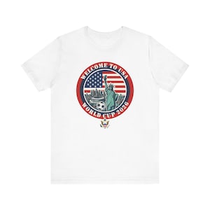 以下が含まれることがあります： 「WELCOME TO USA WORLD CUP 2026」の文字が入った円形グラフィックが特徴の白いTシャツ。自由の女神、サッカーボール、アメリカ国旗がデザインされています。下部には小さなエンブレムがあります。