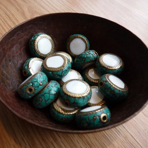Puede incluir: Un cuenco de cuentas ovaladas de color turquesa y blanco con detalles dorados. Las cuentas están hechas de una combinación de materiales, incluyendo turquesa, concha blanca y oro.