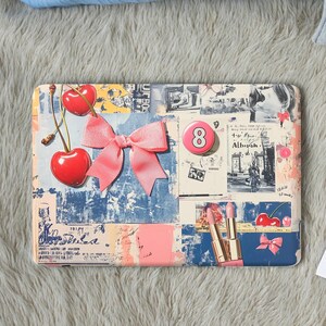 K&ouml;nnte beinhalten: Eine rechteckige Laptop-Folie mit einer Collage aus blauen, pfirsichfarbenen und wei&szlig;en Texturen. Sie zeigt zwei rote Kirschen, eine gro&szlig;e rosa Stoffschleife, einen rosa Knopf mit der Zahl 8, ein Schwarz-Wei&szlig;-Foto und zwei rosa Lippenstifte.