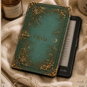 May include: A teal e-reader case with gold filigree corners and edges, featuring the name Olivia and crescent moon symbols in the center. The case includes a hand strap, a foldable stand, and a magnetic closure, designed to protect a black Kindle.