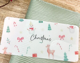 May include: A white rectangular card with a Christmas theme, featuring illustrations of snowmen, deer, Christmas trees, and candy canes. The card is placed on top of a green textured wallet. The word "Christmas" is written in cursive.