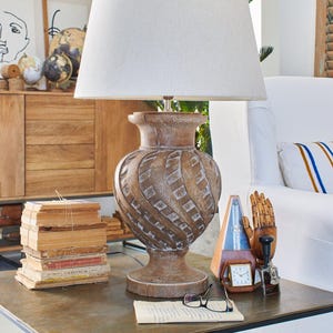 May include: A table lamp with a beige fabric shade and a carved wooden base with a spiral design. The lamp is on a metal table with books, a metronome, and other decorative items. The base is made of wood.