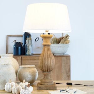Pode incluir: Um abajur de mesa de madeira com um abajur de tecido branco. A lâmpada tem uma base de madeira esculpida e um abajur de tecido branco. A lâmpada está sobre uma mesa de madeira com um livro e óculos em cima.