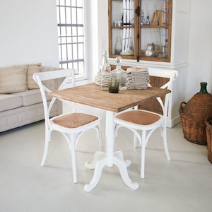 Può includere: Un tavolo da pranzo quadrato bianco con un piano in legno e una base a piedistallo bianca. Due sedie bianche con sedute in canna intrecciata sono posizionate attorno al tavolo. Le sedie hanno un design semplice ed elegante con uno schienale curvo e un supporto a forma di croce.