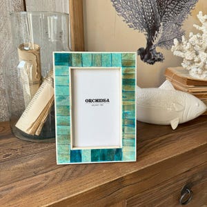 Può includere: Una cornice per foto in mosaico verde acqua e verde con un passepartout bianco e il testo "Orchidéa Milano 1981".