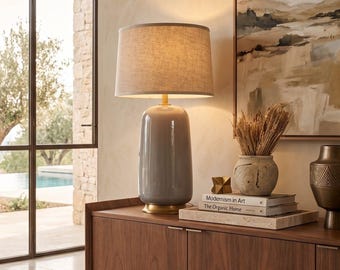Può includere: Una lampada da tavolo con base in ceramica grigia lucida, collo e base circolare in metallo dorato e un paralume cilindrico in tessuto beige. La lampada emette una luce calda, illuminando l'area circostante con un bagliore morbido.