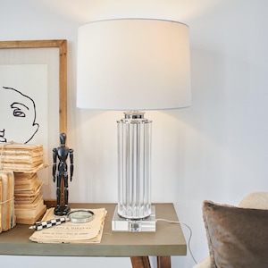 Op de afbeelding: Een moderne tafellamp met een heldere, geribbelde glazen voet en een witte lampenkap met een lichtblauwe rand. De lamp staat op een grijze tafel met andere decoratieve items, waaronder een zwart houten mannequin en een ingelijste schets.