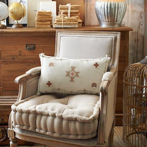 Könnte beinhalten: Ein hellholzfarbener Sessel mit einem beigen Leinenkissen und einem gemusterten Kissen. Der Sessel hat einen gesteppten Sitz und einen dekorativen Holzrahmen. Der Sessel steht vor einer Kommode aus Holz mit einer Lampe darauf.