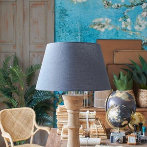 Puede incluir: Una pantalla de lámpara gris con una base de madera y una pila de libros sobre una mesa de madera. La lámpara está delante de un papel tapiz floral azul y blanco.