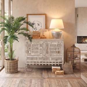 Op de afbeelding: Woonkamer met een witte, verweerde kast, een zilveren lamp en een ingelijste lijntekening. Een palmboom in een gevlochten mand en een decoratieve vogelkooi dragen bij aan de inrichting. De kamer heeft houten vloeren en een open haard.