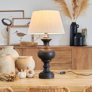 Może przedstawiać: Czarna lampa stołowa z kremowym abażurem stoi na drewnianym stole. Lampa ma okrągłą podstawę i wysokie, smukłe ciało. Stół jest otoczony wiklinowymi krzesłami i ozdobnymi przedmiotami.