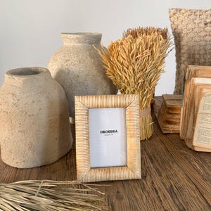 May include: A natural woven picture frame with a white background and the text "Orchidéa Milano 1981". The frame is sitting on a wooden table with dried grasses and ceramic vases.