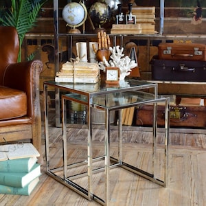 Op de afbeelding: Twee glazen nesttafels met een chroom frame. De tafels staan op een houten vloer.