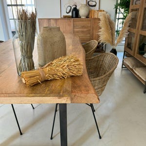 Könnte beinhalten: Ein Holztisch mit schwarzen Metallbeinen ist mit dekorativen Gegenständen bestückt. Eine klare Glasvase enthält getrocknete Gräser, und eine strukturierte Vase und ein Weizenbündel befinden sich auf dem Tisch. Korbstühle und ein Holzschrank sind im Hintergrund.