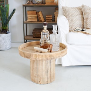 Op de afbeelding: Een ronde, lichtgekleurde houten salontafel met een getextureerde, geribbelde basis. Twee glazen flessen, een karaf en een opgerolde rol liggen erop. Een witte bank en een plank met boeken en globes staan op de achtergrond.