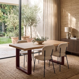 Può includere: Un tavolo da pranzo rettangolare in legno con un piano spesso color marrone chiaro e due gambe in metallo a forma di U color rosso scuro. Quattro sedie con sedute imbottite color crema e sottili telai in metallo nero circondano il tavolo.