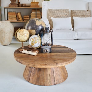 Può includere: Un tavolino rotondo in legno rustico con base a piedistallo. Il tavolo è adornato con mappamondi vintage, una lanterna e una macchina fotografica. Un divano bianco e oggetti decorativi sullo sfondo creano un'atmosfera da soggiorno vintage.
