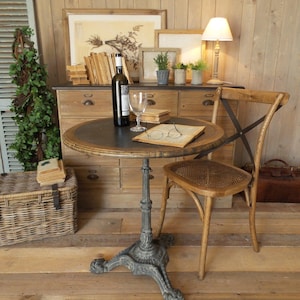 Peut inclure: Une table de bistrot ronde avec un plateau sombre et un pied en fonte orné. Une chaise en bois avec une assise en osier est à côté de la table. Une bouteille de vin, un verre et un livre sont sur la table. Une lampe et des œuvres encadrées sont en arrière-plan.