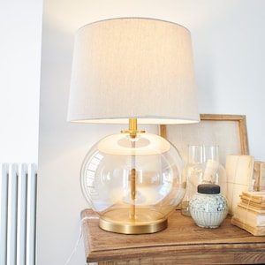 Può includere: Una lampada da tavolo decorativa con una base in vetro trasparente e sferica e un paralume in tessuto beige. La lampada ha una base e dettagli in metallo dorato. È posizionata su un tavolo di legno, con altri oggetti decorativi nelle vicinanze.
