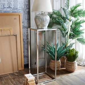 Op de afbeelding: Een zilveren lamp met een witte lampenkap staat op een verchroomde standaard met een glazen blad. De standaard staat naast een plant in een pot met groene bladeren en een rieten mand.