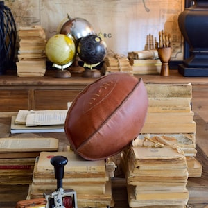 Pode incluir: Uma almofada em forma de bola de futebol de couro castanho repousa sobre uma superfície de madeira, rodeada por pilhas de livros antigos. Um relógio vintage, um carimbo e globos também são visíveis, criando uma estética clássica e erudita.