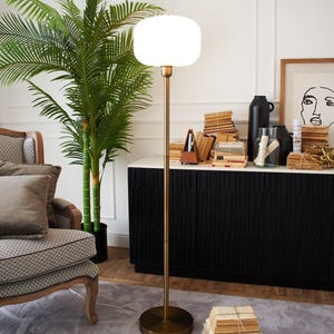 Peut inclure: Un abat-jour de lampe rond et blanc en verre dépoli avec une base en métal doré et une tige longue et fine. La lampe est posée sur un tapis gris devant un meuble noir.