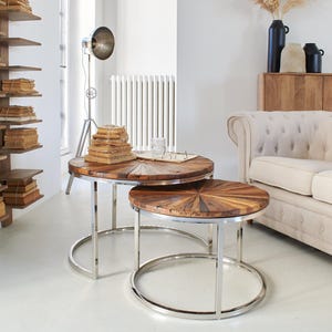 Op de afbeelding: Twee ronde houten salontafels met een chromen onderstel. De bovenste tafel heeft een zonnestralenontwerp en de onderste tafel is iets kleiner.