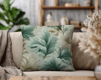 May include: Square throw pillow with a botanical design. The pillow features large, stylized leaves in shades of green and cream against a light background. The pillow is placed on a beige sofa, suggesting a home decor theme.