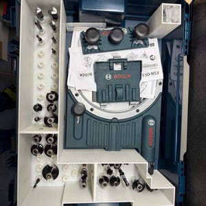 May include: An open, dark teal Bosch tool case filled with various router bits and accessories. The case has white storage compartments and includes a Bosch router and instruction manuals. The Bosch logo is visible on the case and tools.