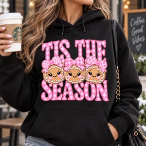 Può includere: Una felpa nera con cappuccio caratterizzata da una stampa grafica di tre biscotti di pan di zenzero con fiocchi rosa. Il testo TIS THE SEASON è scritto in grandi lettere rosa con un motivo a fiocchi di neve bianchi.