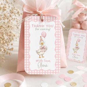 May include: A rectangular gift tag with a pink and white gingham border featuring a cartoon duck wearing a party hat and holding a pink balloon. The text reads THANK YOU for coming! and With love, Olivia. The tag is tied to a pink favor bag.