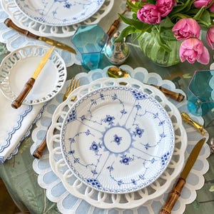 Pode incluir: Uma mesa posta com pratos brancos com desenhos florais azuis,  placemats de renda branca, talheres de ouro e um vaso de tulipas cor-de-rosa.