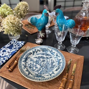Puede incluir: Una mesa puesta con platos de porcelana azul y blanca, cubiertos dorados y dos figuras de gallo turquesa. Los platos presentan un detallado diseño floral azul. También hay copas de vino transparentes y una jarra.