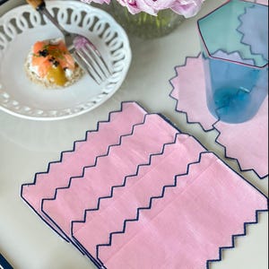 Può includere: Tovaglioli di lino rosa con bordo a festoni e bordatura blu scuro. I tovaglioli sono impilati su una superficie bianca.