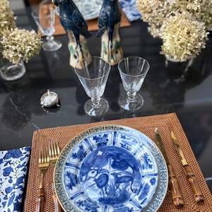 Puede incluir: Una elegante mesa con platos de porcelana azul y blanca, cubiertos dorados con mangos de bambú y copas de vino transparentes. Dos figuras de pájaros azules adornan la mesa. Los platos presentan un diseño detallado de animales y flores, sobre un mantel individual tejido.