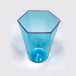 Pode incluir: Um vaso de vidro transparente em forma hexagonal, com um tom azul claro. A borda é delineada com uma fina borda vermelha. O vaso tem um design alargado, mais largo no topo e afinando em direção à base. A parte inferior do vaso tem um design em espiral.