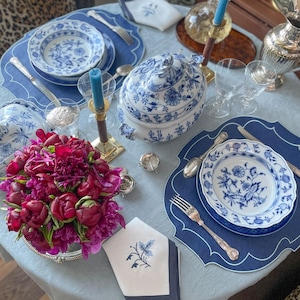 Pode incluir: Uma mesa posta com louça de padrão azul e branco, uma sopeira, pratos e guardanapos. A mesa está posta com uma toalha de mesa azul e  placemats azuis. Também há flores em um vaso de prata.