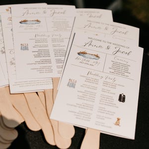 May include: Wedding ceremony program fans with a watercolor illustration of a lake and the text "Welcome to the wedding of Anna & Jacob" printed on the front. The back of the fan lists the wedding party and the ceremony order.