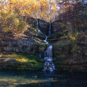 Puede incluir: Una cascada cae sobre rocas cubiertas de musgo en un exuberante entorno forestal. El agua es clara y las rocas son una mezcla de marr&oacute;n y gris. Las hojas de los &aacute;rboles son una mezcla de verde y amarillo, creando una hermosa escena de oto&ntilde;o.