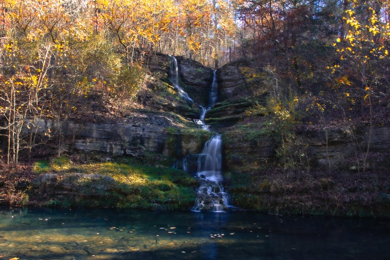 Puede incluir: Una cascada cae sobre rocas cubiertas de musgo en un exuberante entorno forestal. El agua es clara y las rocas son una mezcla de marr&oacute;n y gris. Las hojas de los &aacute;rboles son una mezcla de verde y amarillo, creando una hermosa escena de oto&ntilde;o.