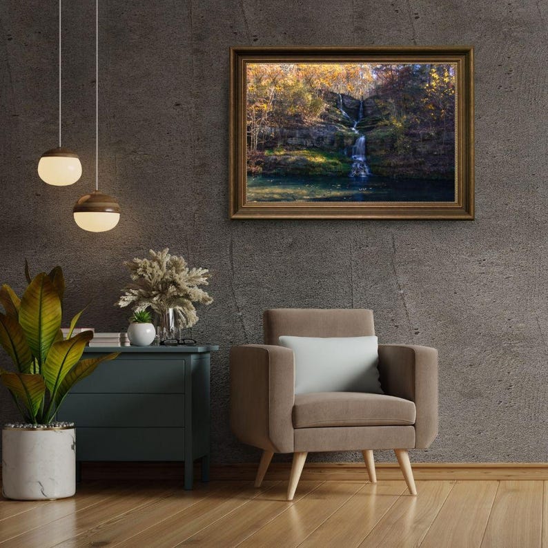 Puede incluir: Una fotograf&iacute;a enmarcada de una cascada que cae por un acantilado rocoso, rodeada de exuberante vegetaci&oacute;n verde y hojas de oto&ntilde;o. La fotograf&iacute;a est&aacute; colgada en una pared gris en un ambiente de sala de estar, con un sill&oacute;n marr&oacute;n y una mesa auxiliar con una planta en maceta.